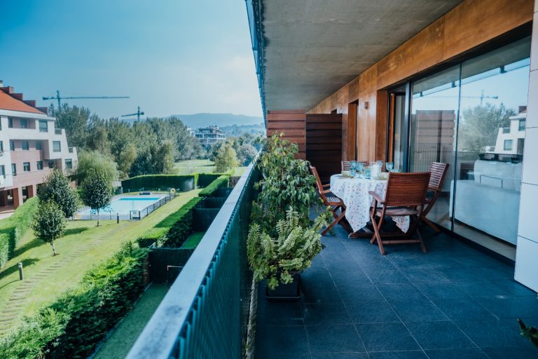 Espacioso balcón de un apartamento con suelo de baldosas oscuras y una vista panorámica de un área residencial verde. El balcón está amueblado con una mesa de madera y sillas plegables, con una mantelería estampada y una mesa preparada para una comida al aire libre.
